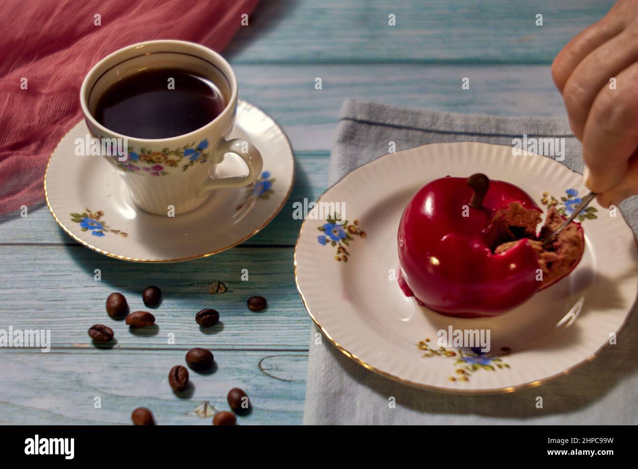 Frühstückskonzept mit сherry-Kuchen auf Keramikplatte, Kaffee in einer Fayence-Tasse, verstreuten Kaffeebohnen und einer menschlichen Hand, die einen Kuchen nimmt. Stockfoto