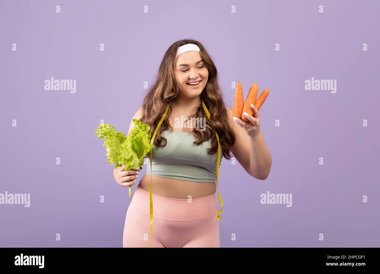 Glückliche plus Größe kaukasische junge Frau in Sportbekleidung mit Maßband Wahl Karotten oder Salatblätter, isoliert auf violettem Hintergrund, Studio. Wei Stockfoto