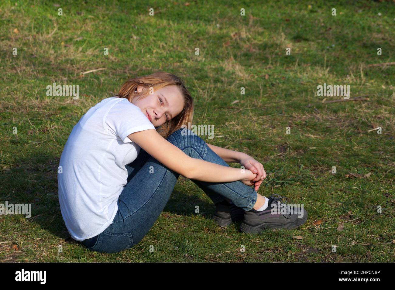 Lächelndes Teenager-Mädchen in einem weißen T-Shirt sitzt auf dem Gras mit dem Kopf auf den Knien. Stockfoto