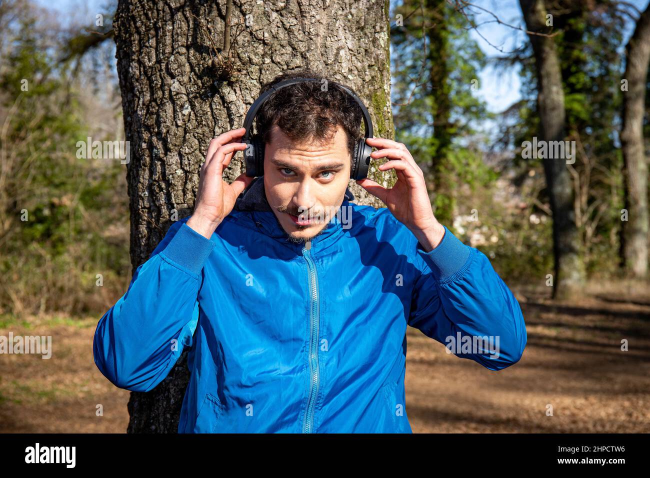 Porträt eines hübschen jungen Mannes mit blauen Augen und einem Schnurrbart, der beim Training Musik hört. Der Mann in der blauen Jacke trainiert in t Stockfoto