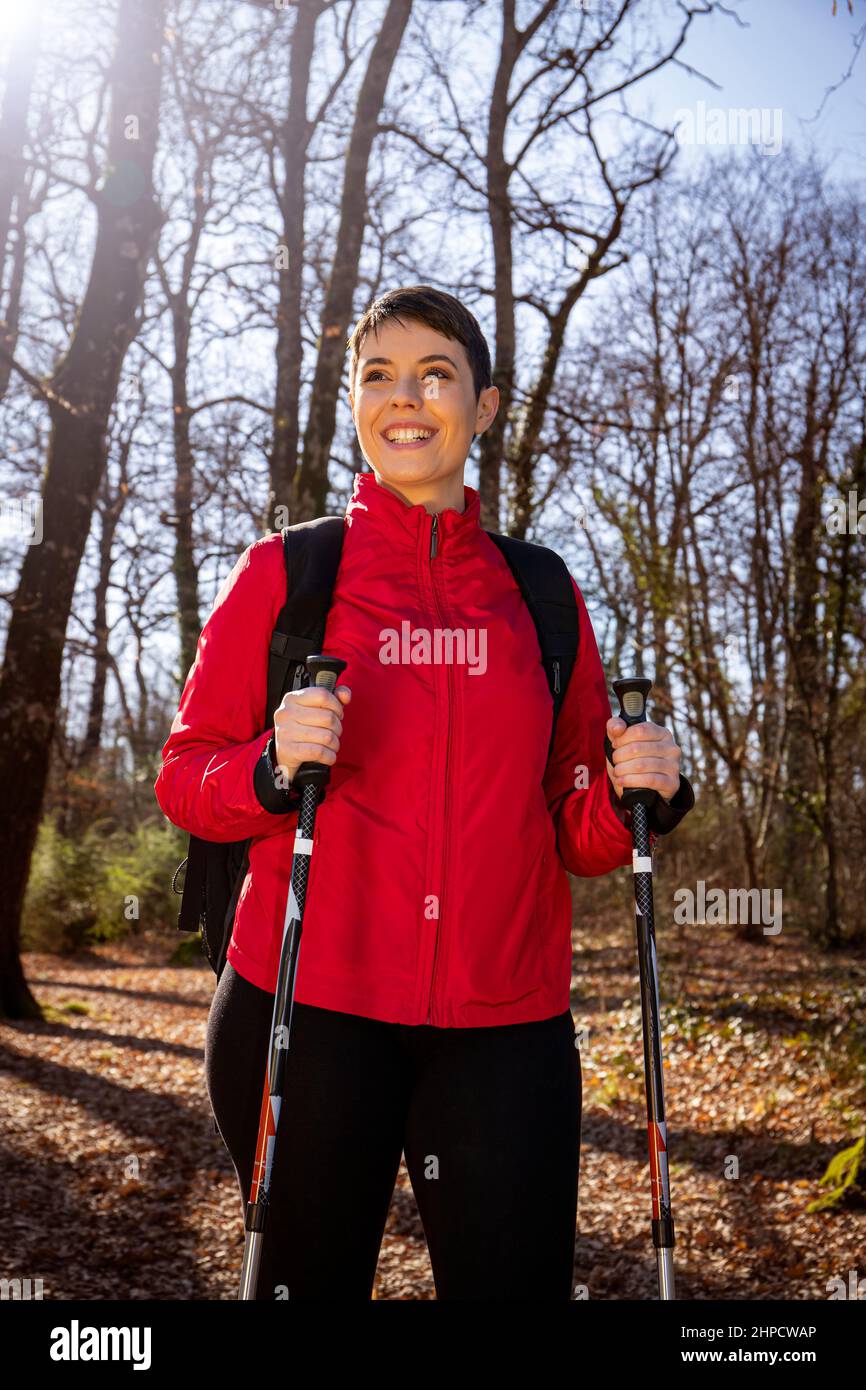 Die lächelnde junge Frau wandert im Wald. Die Frau hat kurze Haare, trägt eine rote Jacke und verwendet Trekkingstöcke. Reise- und Outdoor-Konzept. Stockfoto