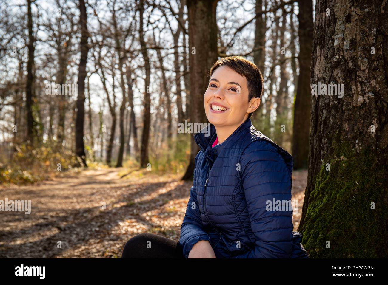 Eine junge lächelnde Frau sitzt am Fuße einer Eiche im Wald. Schöne Frau ruht sich nach einem Spaziergang im Park aus. Reise- und Outdoor-Konzept. Stockfoto