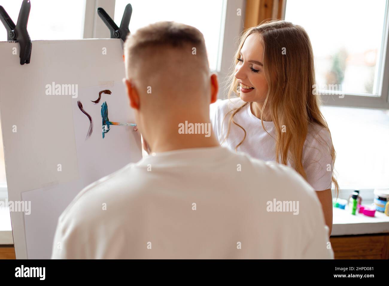 Rückansicht des Mannes stehen in der Nähe junge nette lächelnde Frau mit langen hellen Haaren in weißem T-Shirt Malerei absorbierenden auf Leinwand. Stockfoto