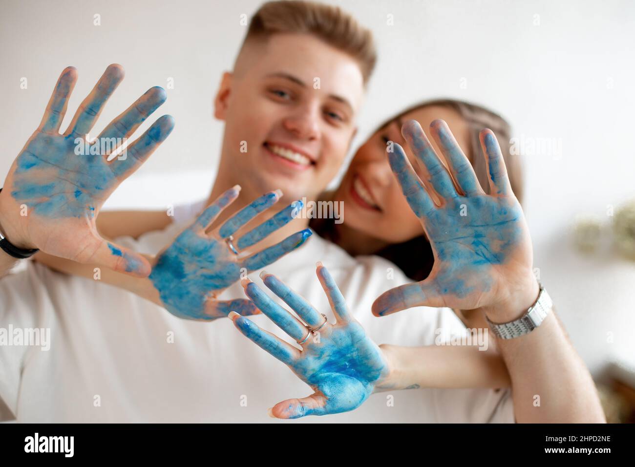 Glücklich lächelndes junges Paar hübscher Frau steht hinter dem Mann in weißen T-Shirts und zeigt Hände, die mit blauer Farbe bedeckt sind. Stockfoto