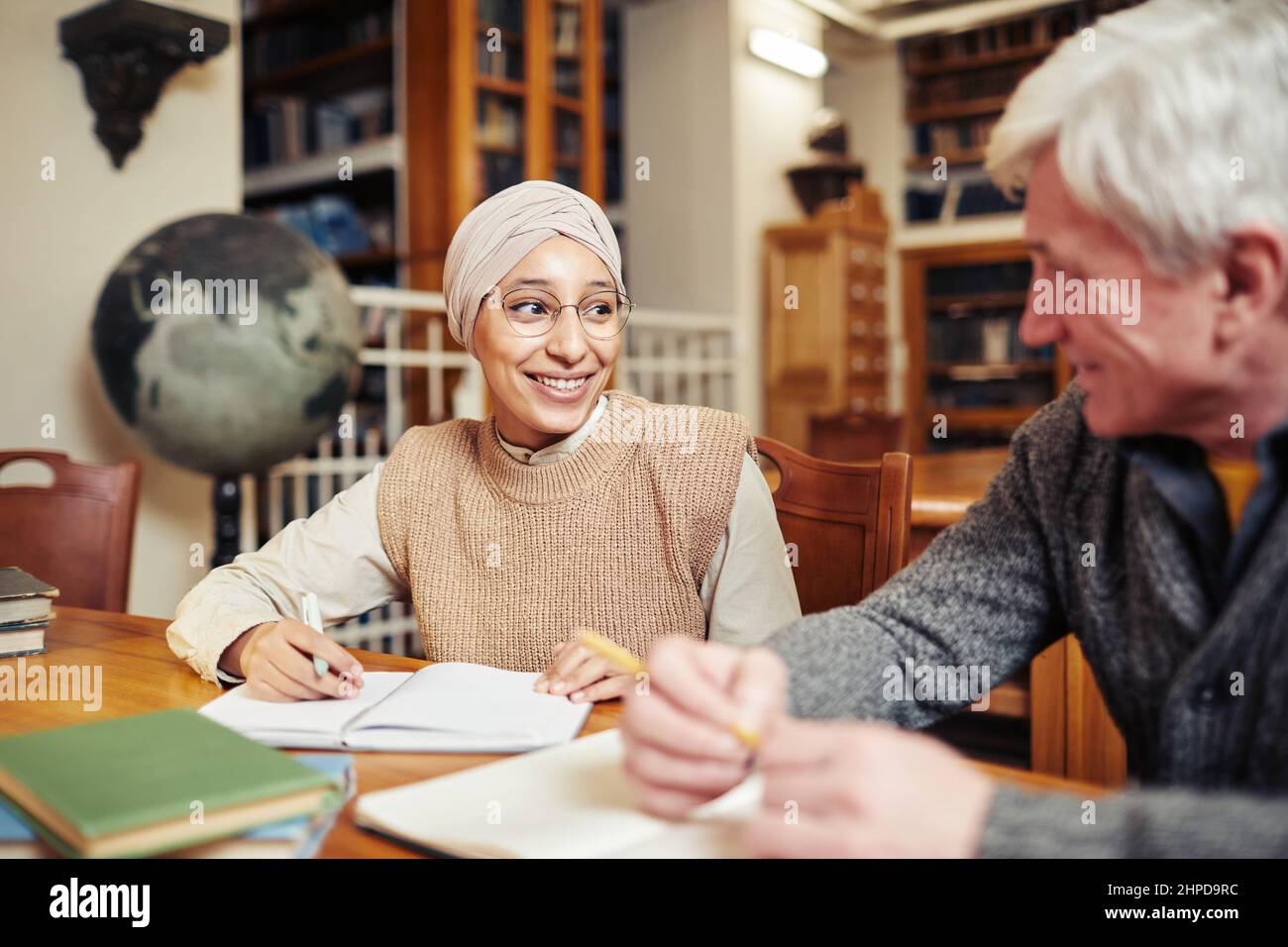 Porträt einer lächelnden muslimischen Frau mit Kopfbedeckung während des Studiums in der Universitätsbibliothek bei einem Senior Professor Stockfoto