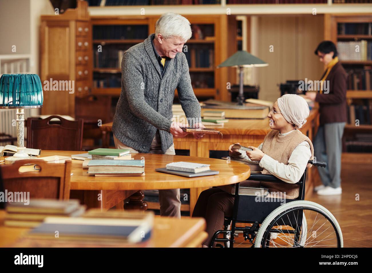 Porträt eines lächelnden Seniorprofessors, der jungen Frau im Rollstuhl in der Universitätsbibliothek hilft Stockfoto