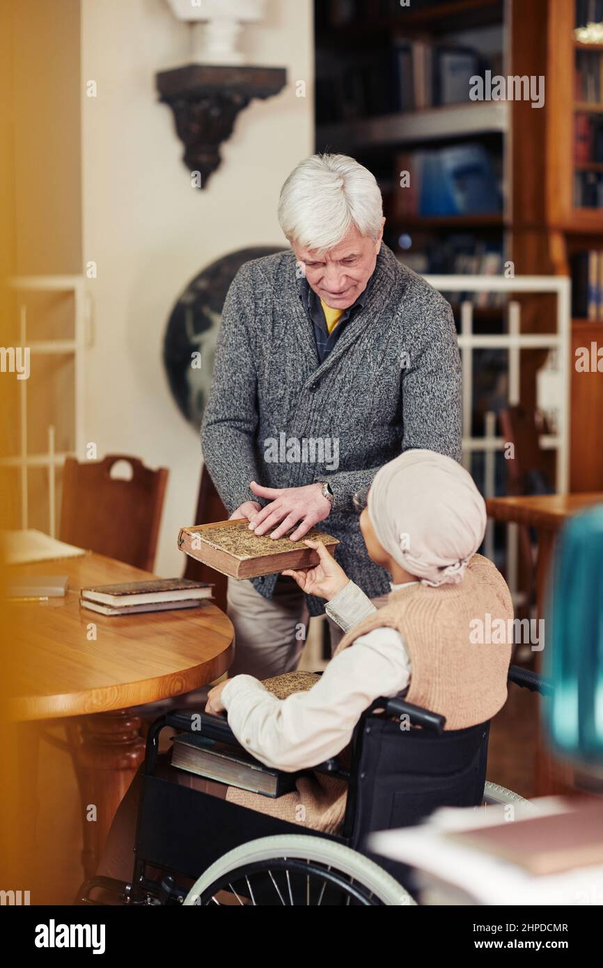 Vertikales Porträt eines lächelnden Seniorprofessors, der jungen Frau im Rollstuhl in der Universitätsbibliothek hilft Stockfoto