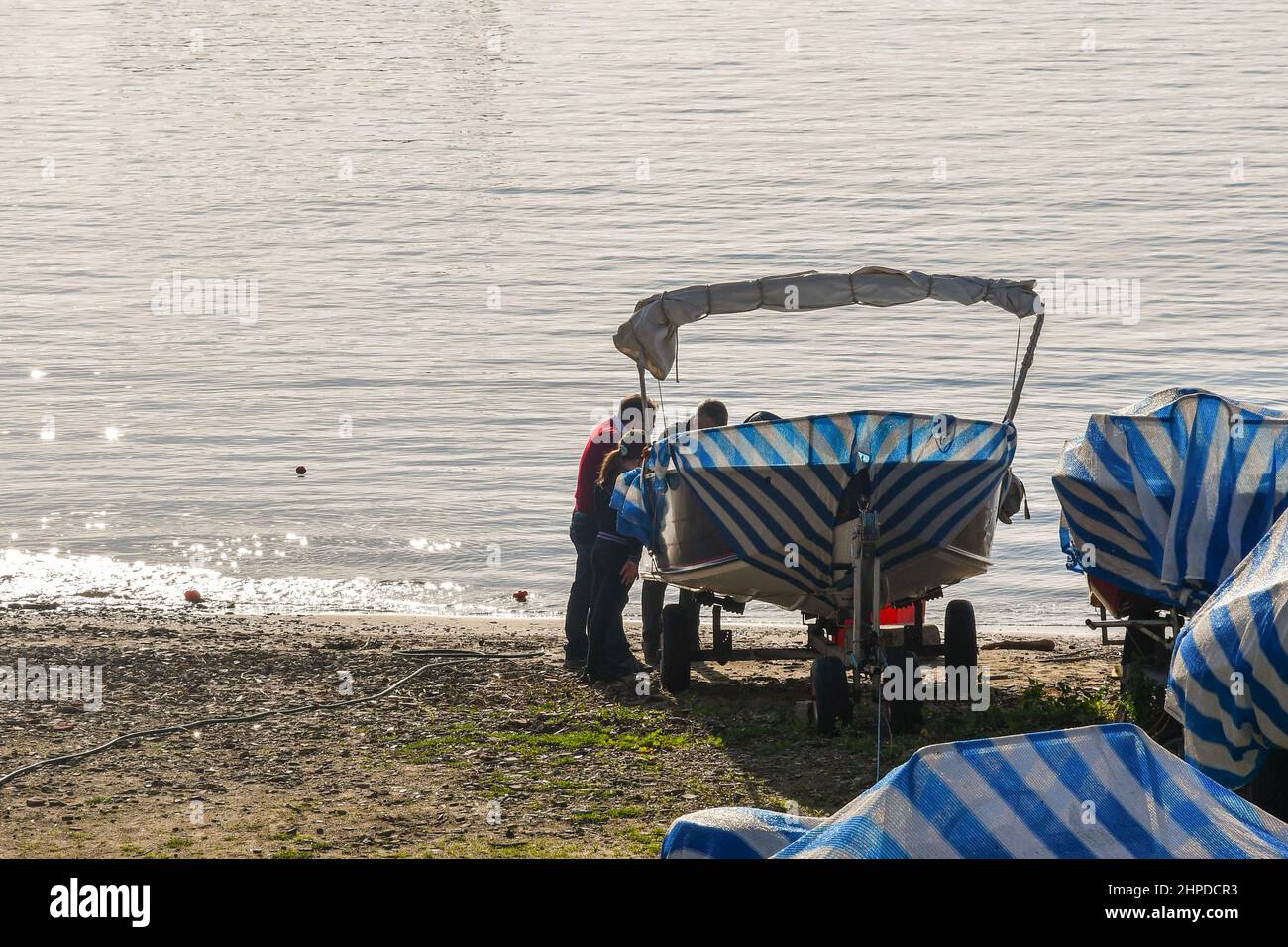 Blick auf den Strand mit Blick auf die Hintergrundbeleuchtung, wo die Leute Wartungsarbeiten an einem kleinen Boot in einem Bereich für Winterlager, Sanremo, Imperia, Ligurien, Italien, durchführen Stockfoto