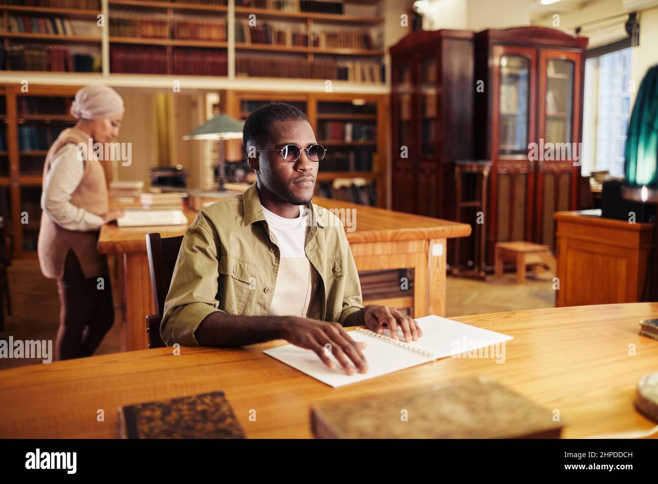 Porträt eines jungen schwarzen Mannes mit Sehbehinderung beim Lesen eines Buches in Braille am Tisch in der Universitätsbibliothek Stockfoto