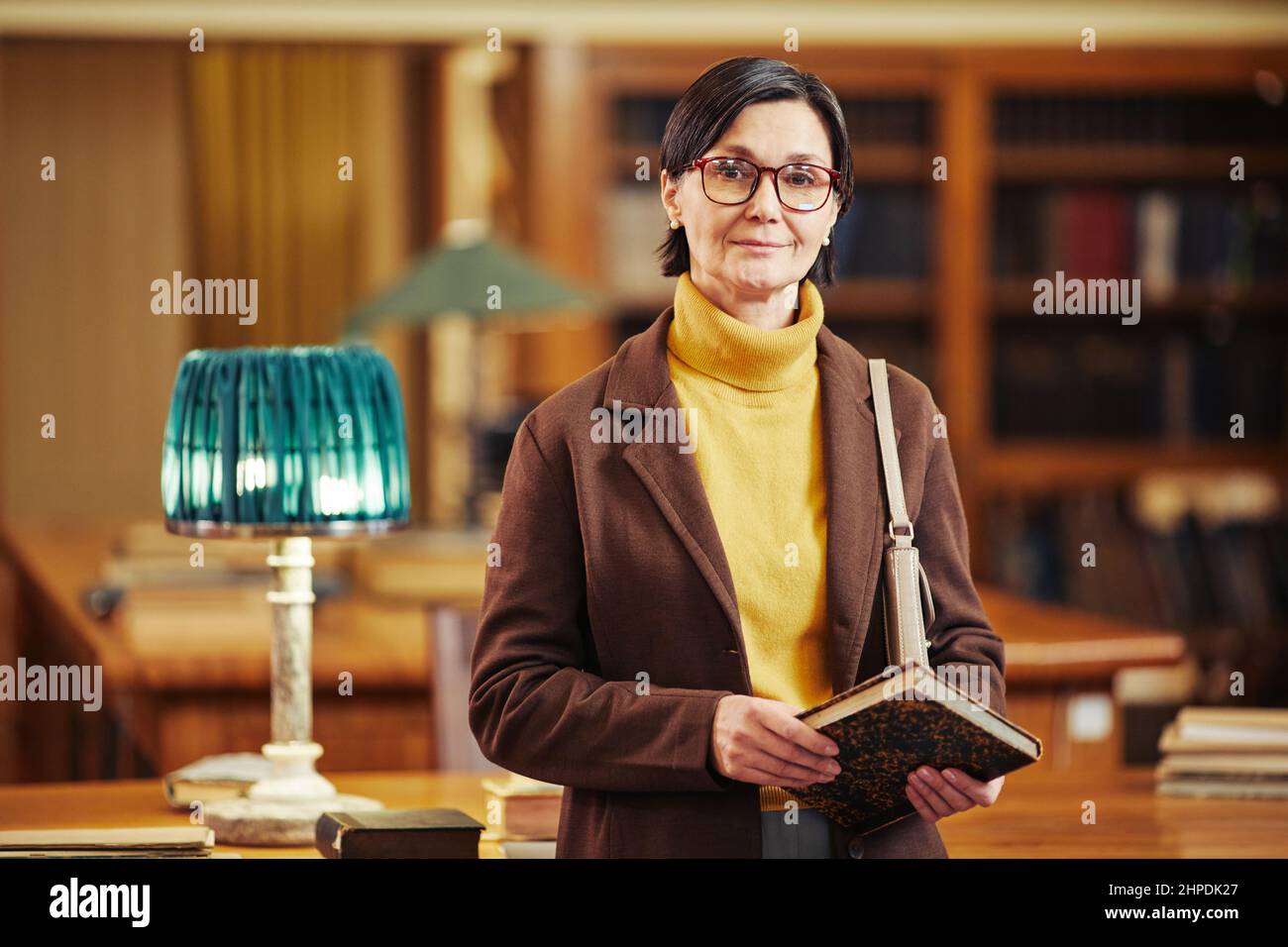 Porträt einer eleganten erwachsenen Frau, die Bücher im Inneren der Bibliothek hält und die Kamera anschaut Stockfoto