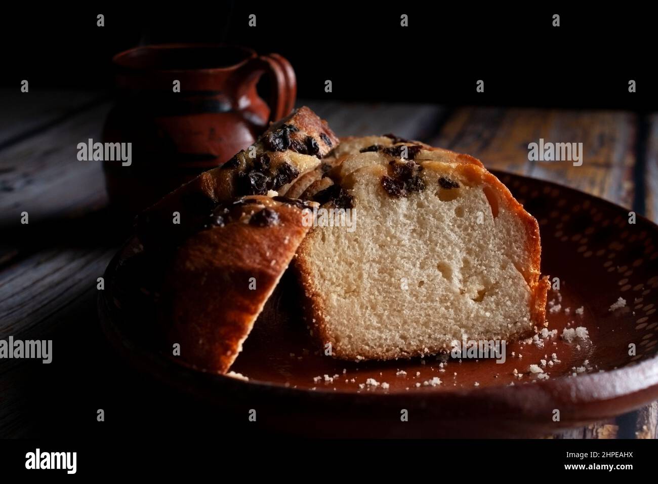 Ein handgebastelter mexikanischer Tonteller mit süßem Brot und Rosinen auf der Oberseite und einer dampfenden Tasse Kaffee oder heißem Schokoladenkakao im Hintergrund. Stockfoto