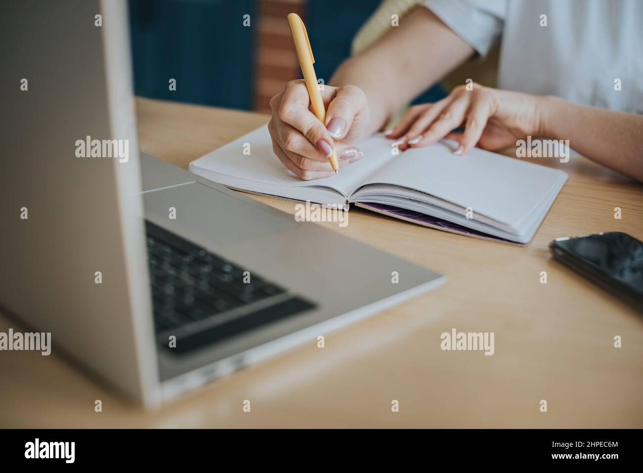 Eine junge Frau sitzt an einem Laptop und schreibt mit einem gelben Stift Notizen in ein Notizbuch. Stockfoto