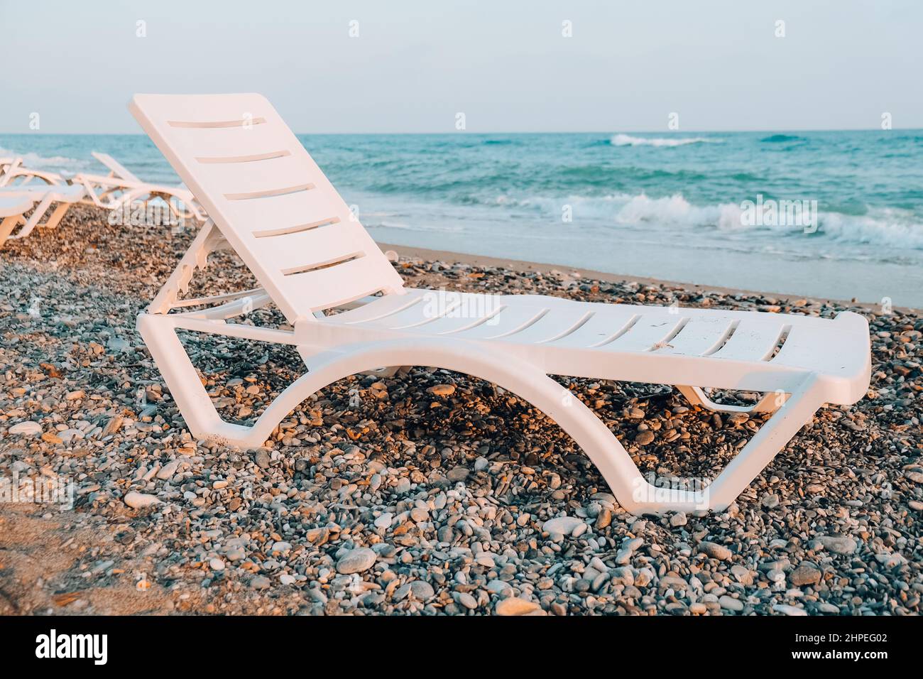 Leere Sonnenliegen vor dem Meer. Liegestühle am Sandstrand. Große Wellen stürmisches Wetter. Eine leere Chaiselongue und ein gefalteter Regenschirm stehen am Rand Stockfoto