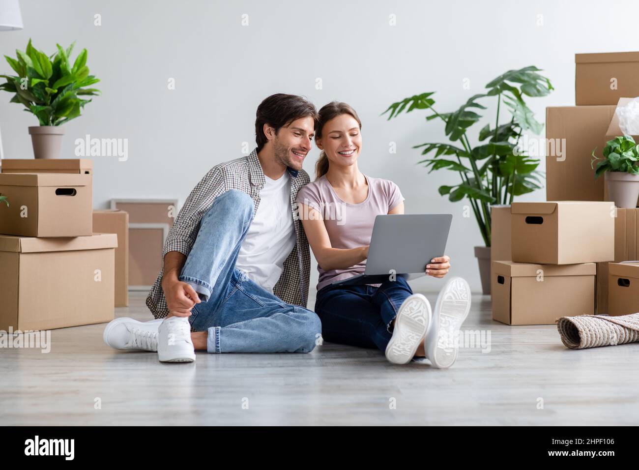 Glad Millennial Caucasian Familie in lässig auf der Suche nach Ideen für neue Innenausstattung auf Computer, sitzt zwischen Pappkartons Stockfoto