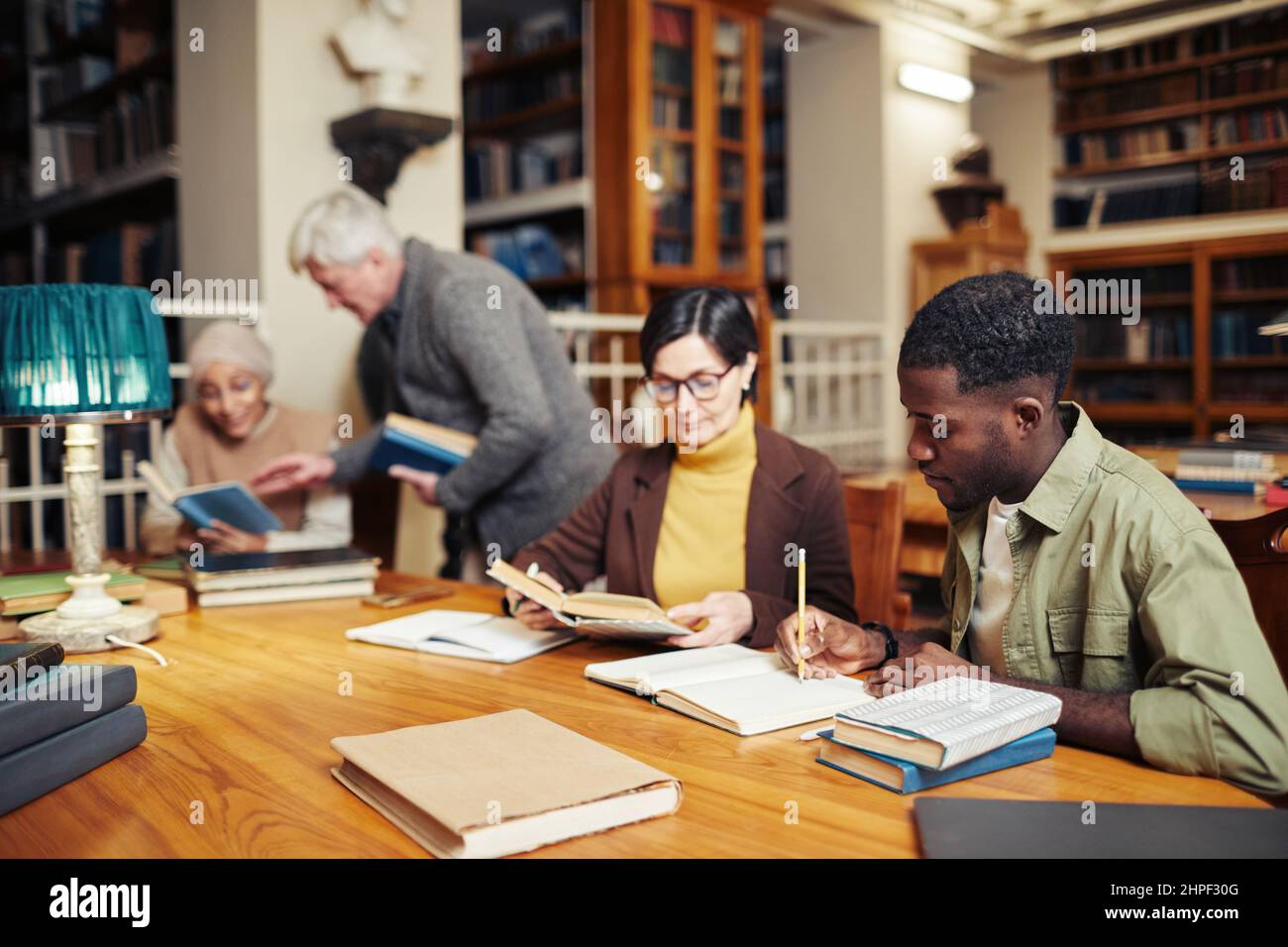Verschiedene Gruppe von Menschen sitzen in Reihe am Tisch in der College-Bibliothek und studieren, konzentrieren sich auf junge schwarze Mann Buch im Vordergrund zu lesen Stockfoto