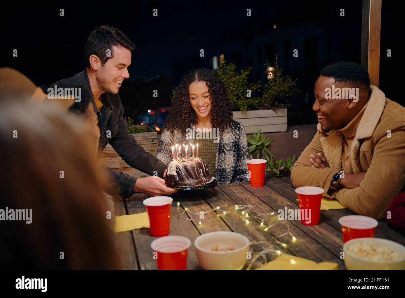 Kaukasischer Mann präsentiert Kuchen für biracial Geburtstagskind. Eine Gruppe von jungen Erwachsenen, die auf der Dachparty am Tisch sitzen und Geburtstag feiern Stockfoto