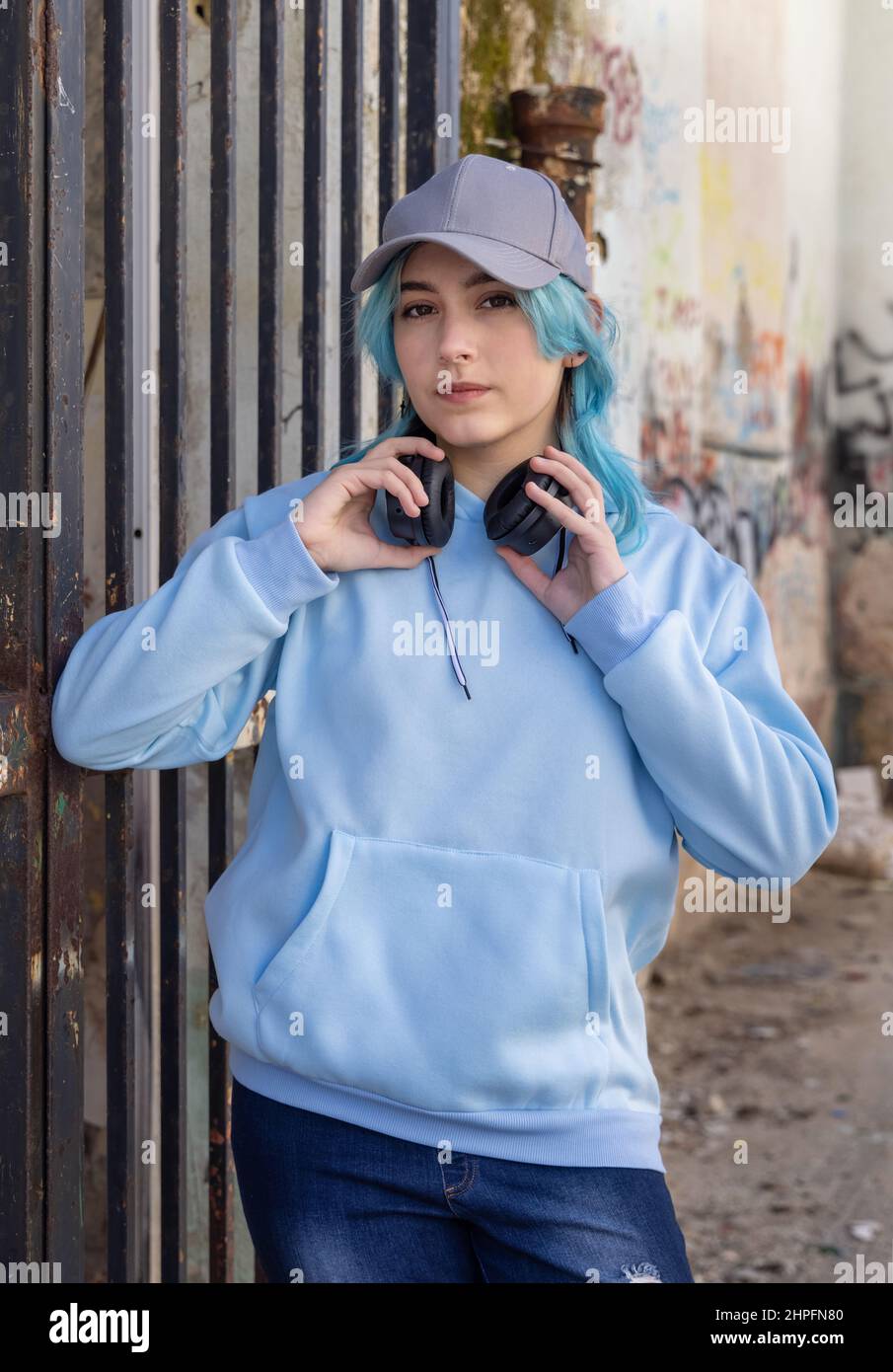Teenager mit hellblauem Oversize Hoodie und Baseballmütze, der in die Kamera schaut. Blauhaarige Teenager-Mädchen mit kabellosen Kopfhörern bleibt im Freien in der Nähe eines Stockfoto
