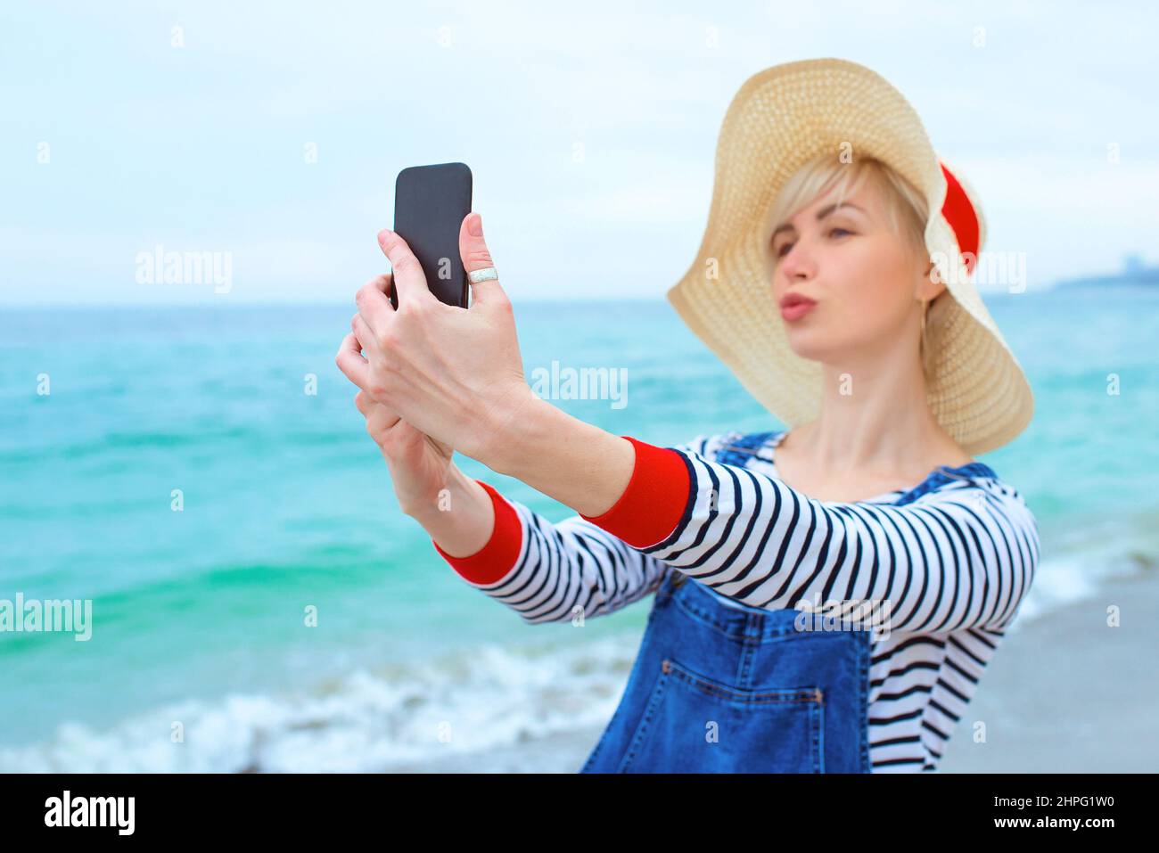 Schöne junge blonde kaukasische Frau im Urlaub in Strohhut, gestreifte Bluse nad Denim Overalls, die selfie auf dem Smartphone am erstaunlichen blauen Meer macht Stockfoto