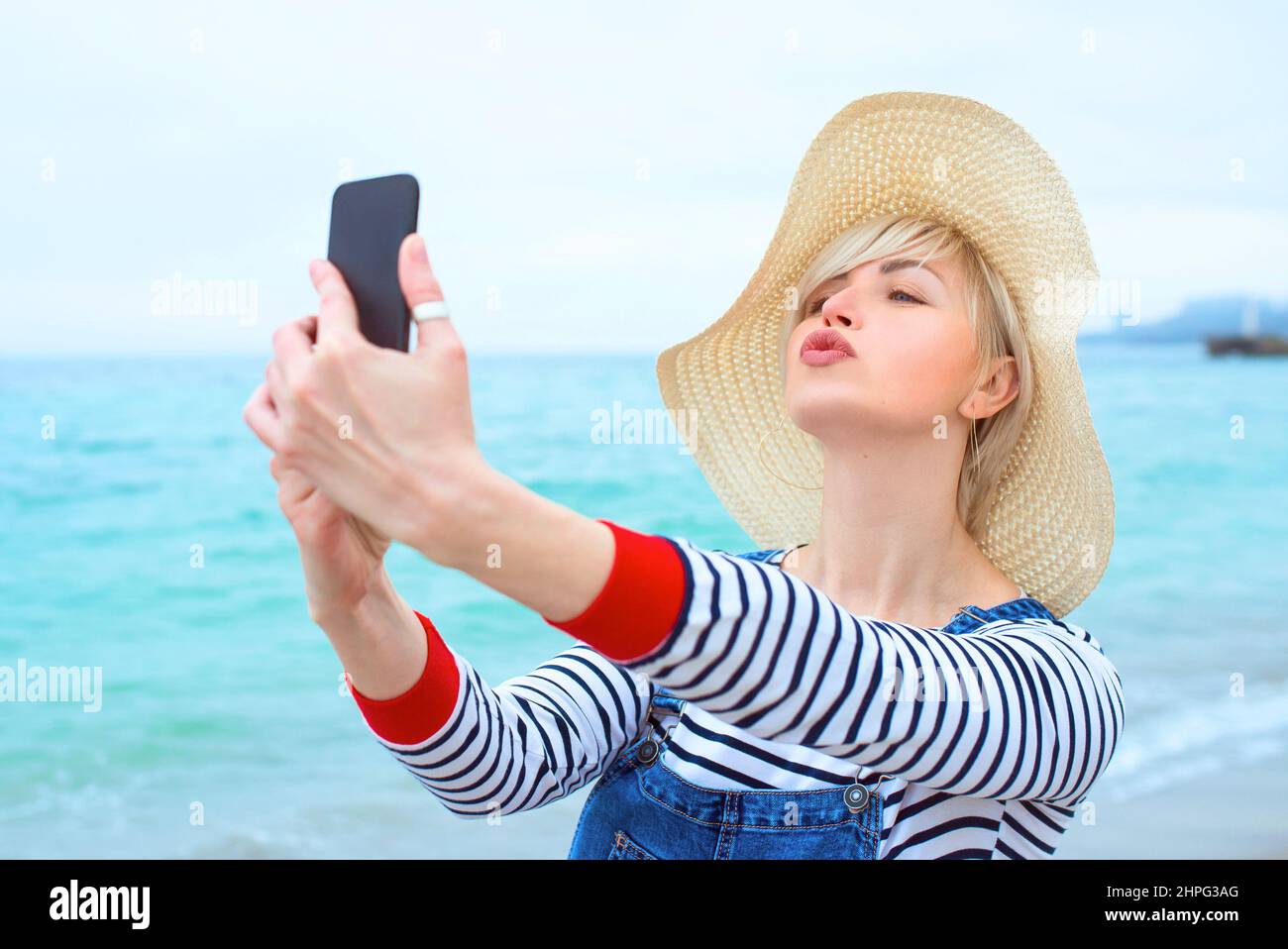 Schöne junge blonde kaukasische Frau im Urlaub in Strohhut, gestreifte Bluse nad Denim Overalls, die selfie auf dem Smartphone am erstaunlichen blauen Meer macht Stockfoto