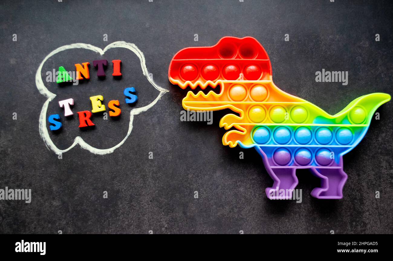 Toy Pop it Dinosaurier Regenbogenfarben auf schwarzem Hintergrund mit bunten Buchstaben und der Aufschrift-Anti-Stress in einer Sprechblase. Stockfoto