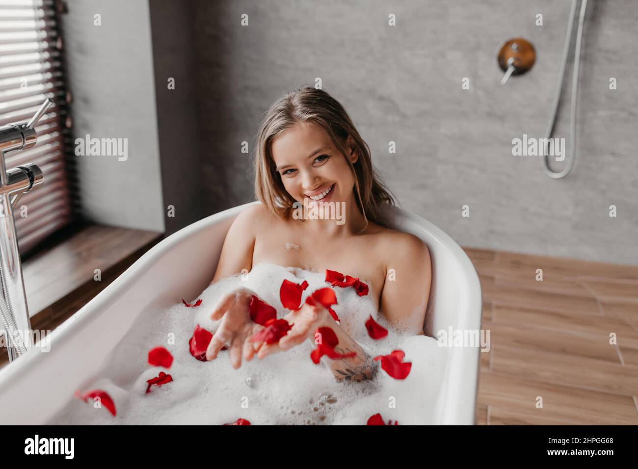 Spa-Behandlungen zu Hause. Fröhliche junge kaukasische Dame, die sich in der Badewanne mit Schaum und Blütenblättern entspannt und bei Tageslicht heißes Wasser im Badezimmerinneren genießt, schauen Sie Stockfoto