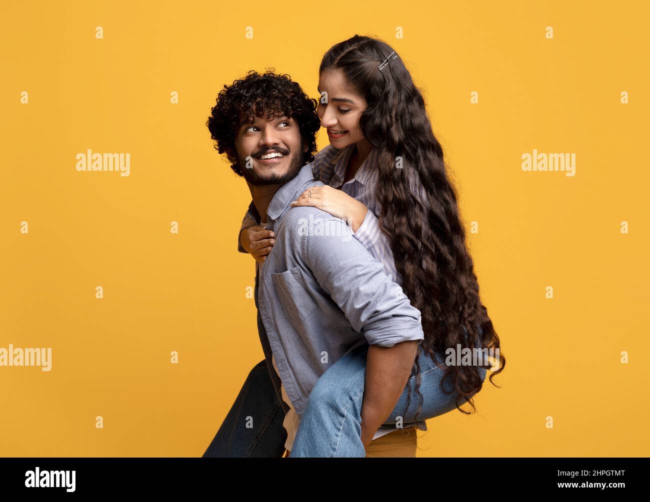 Hübsche indische Dame huckepack ihren Freund, lächelnder Kerl trägt seine Freundin auf dem Rücken, gelber Hintergrund Stockfoto