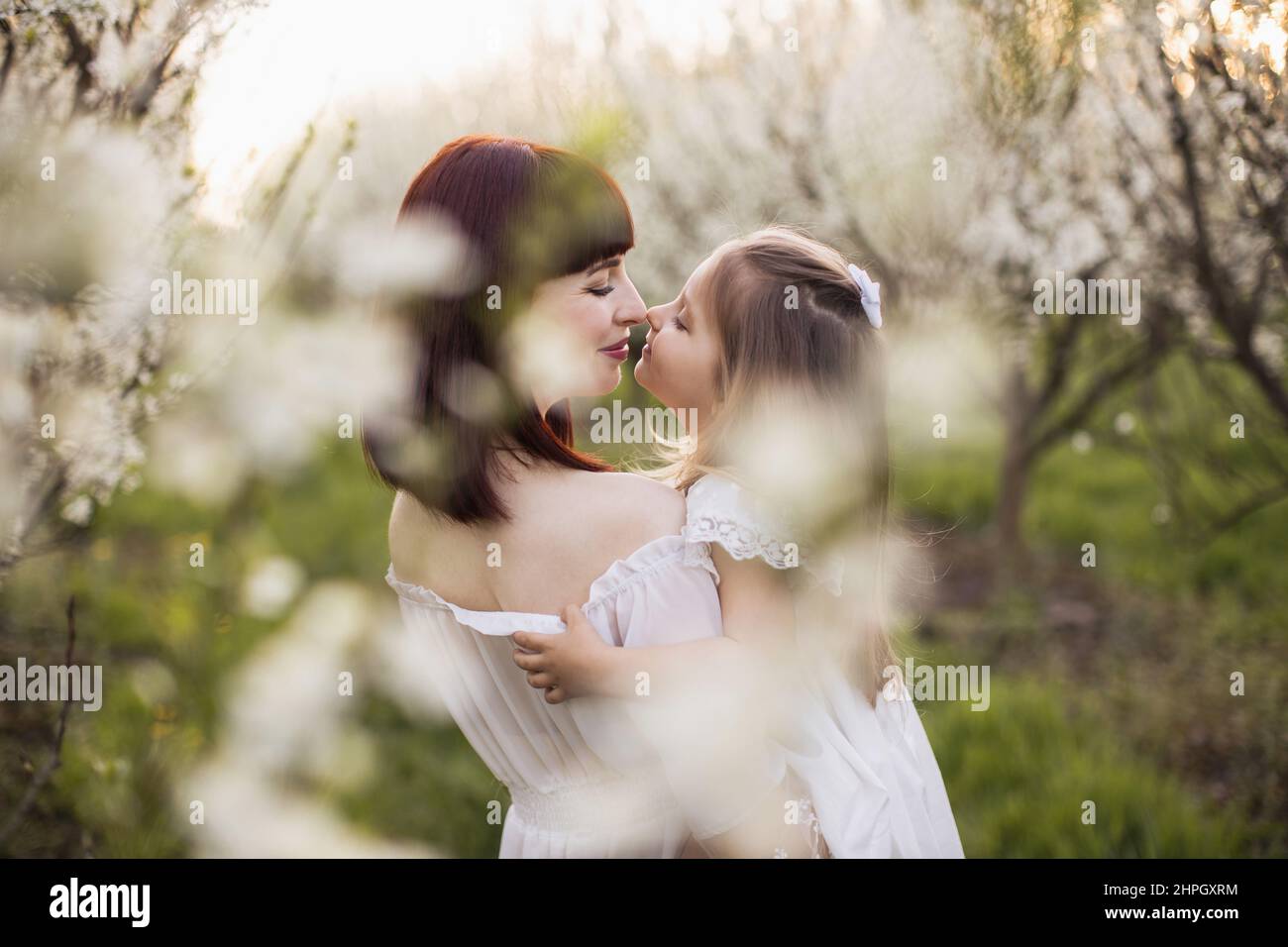 Attraktive junge Frau und kleines weibliches Kind lächeln, während sie mit Nasen im Frühlingsgarten berühren. Mutter und Tochter genießen jeden Moment gemeinsam. Stockfoto