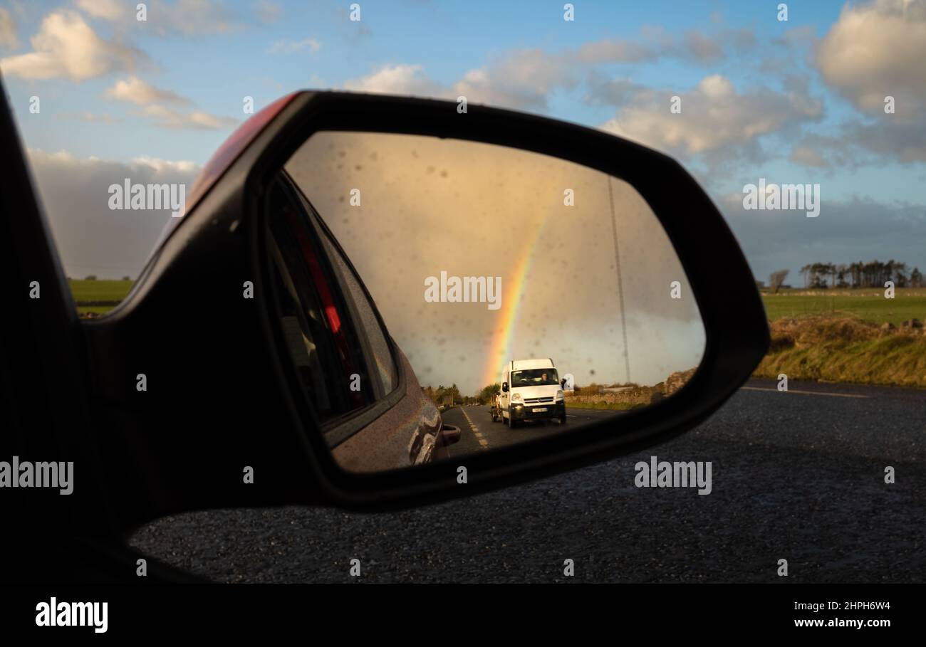 Ein weißer Lieferwagen und ein Regenbogen sind im Seitenspiegel eines Autos zu sehen, das in Irland fährt. Stockfoto