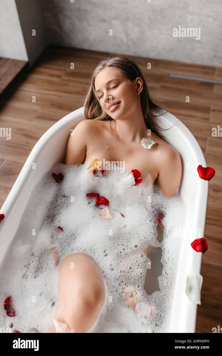 Dame mit geschlossenen Augen des Vergnügens, die zu Hause mit Schaum und Blütenblättern baden und warmes Wasser in der Badewanne genießen Stockfoto