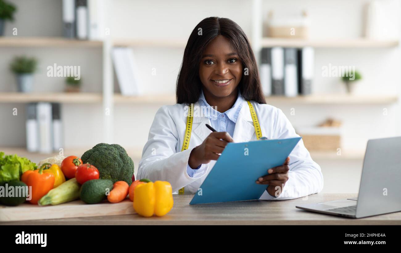 Positive weibliche afroamerikanische Ernährungsberaterin mit Clipboard Erstellung pflanzenbasierte Mahlzeit Plan für Patienten, Panorama Stockfoto