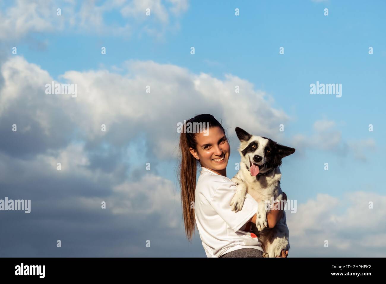 Eine junge lateinische Frau, die ihren Hundefreund in den Armen hält, beide schauen in die Kamera, da ist ein wolkiger blauer Himmel im Hintergrund Stockfoto