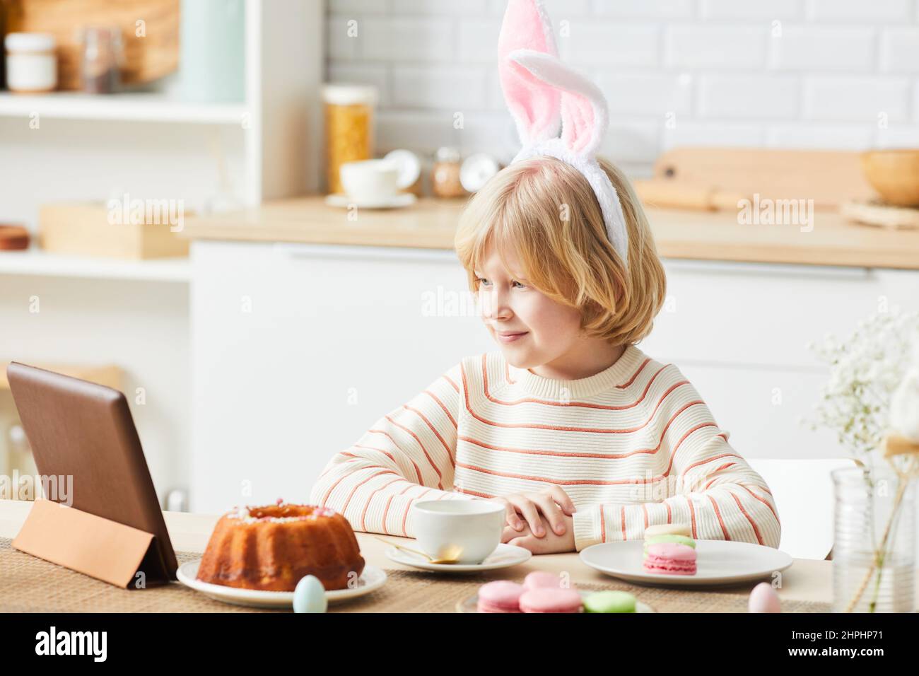 Seitenansicht Porträt eines niedlichen Jungen mit Hasenohren beim Frühstück am Ostermorgen und mit Tablet in der Küche Stockfoto