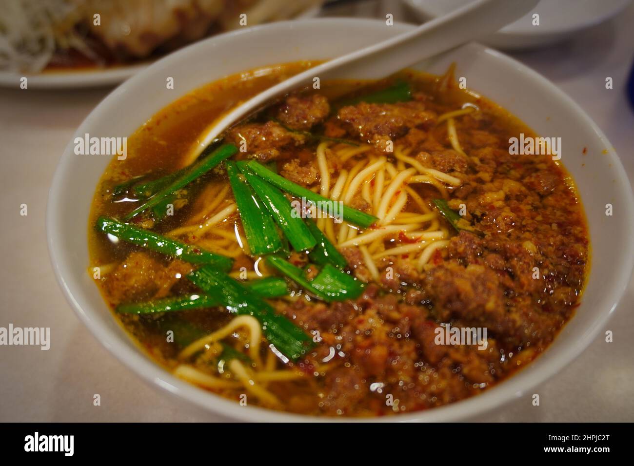 Oben eine Nahaufnahme einer Tasse chinesischer Nudeln, die in einer dicken Fleischbrühe gekocht und in einer weißen Porzellanschüssel mit einer Garnierung mit grünen Zwiebeln serviert werden. Stockfoto