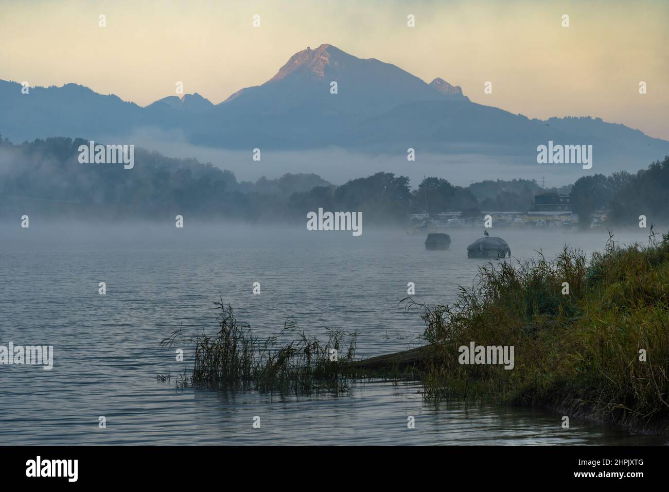 Ufer eines Sees am frühen Morgen des Herbstes. Der Nebel lichtet sich und erlaubt Einblicke in festfahrende Boote. Das Wasser des Sees ist rauh und ein Berg mit seinem Gipfel, der vom ersten Licht beleuchtet wird, ist zu sehen Stockfoto