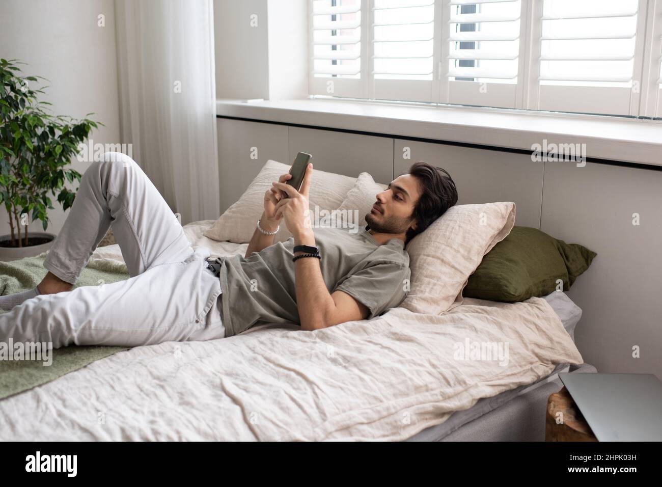 Moderner junger Manager oder Geschäftsmann in Casualwear, der während der Arbeit von zu Hause aus auf dem Bett liegt und per SMS auf dem Smartphone schreibt Stockfoto