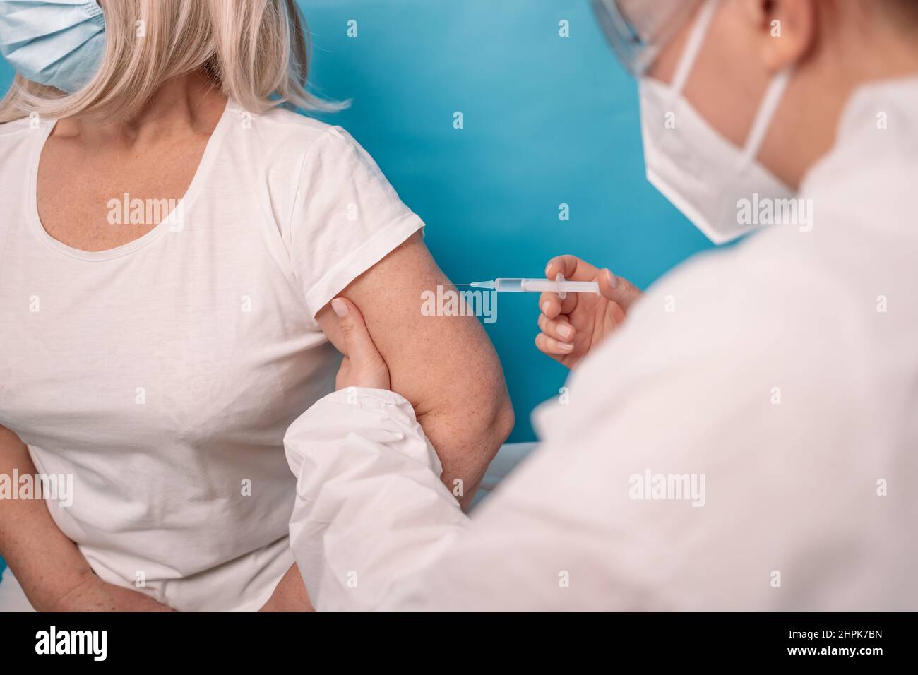 Impfung älterer Menschen. Der Arzt macht eine Impfung gegen ältere Frauen, die Gesichtsmaske auf blauem Hintergrund trägt. Stockfoto