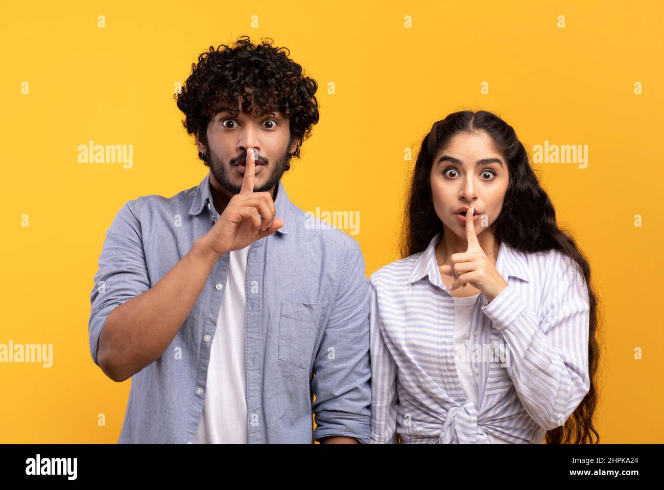 Sagen Sie kein Wort. Indischer Mann und Frau halten die Finger auf den Lippen, machen ein Schweigezeichen, schauen auf die Kamera, gelber Hintergrund Stockfoto