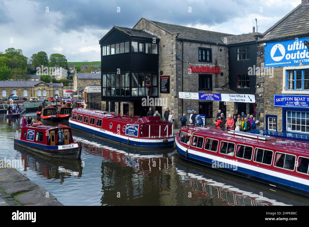 Touristische Freizeitwasserreise (selbstfahrende rote Leihboote, Männer, Frauen, die Schlange stehen, Boote festmachen, Anlegestellen) - landschaftlich schöner Leeds-Liverpool Canal, Yorkshire, England Stockfoto