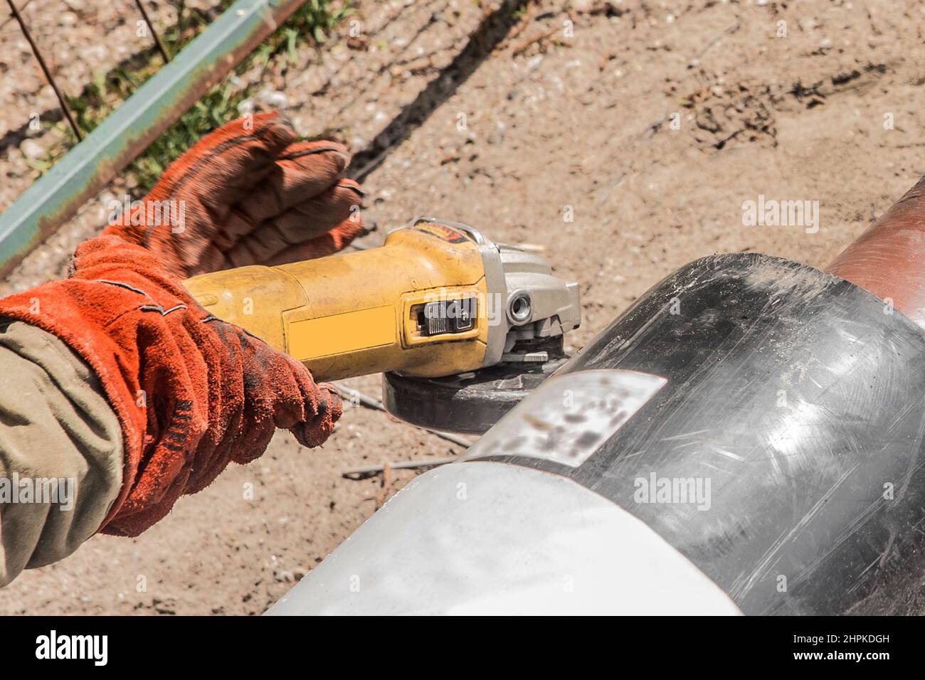 Die Hände eines Industriearbeiters in Arbeitshandschuhe verwenden ein Elektroschleifgerät an einem Polyethylen-Wasserrohr auf einer Baustelle. Stockfoto