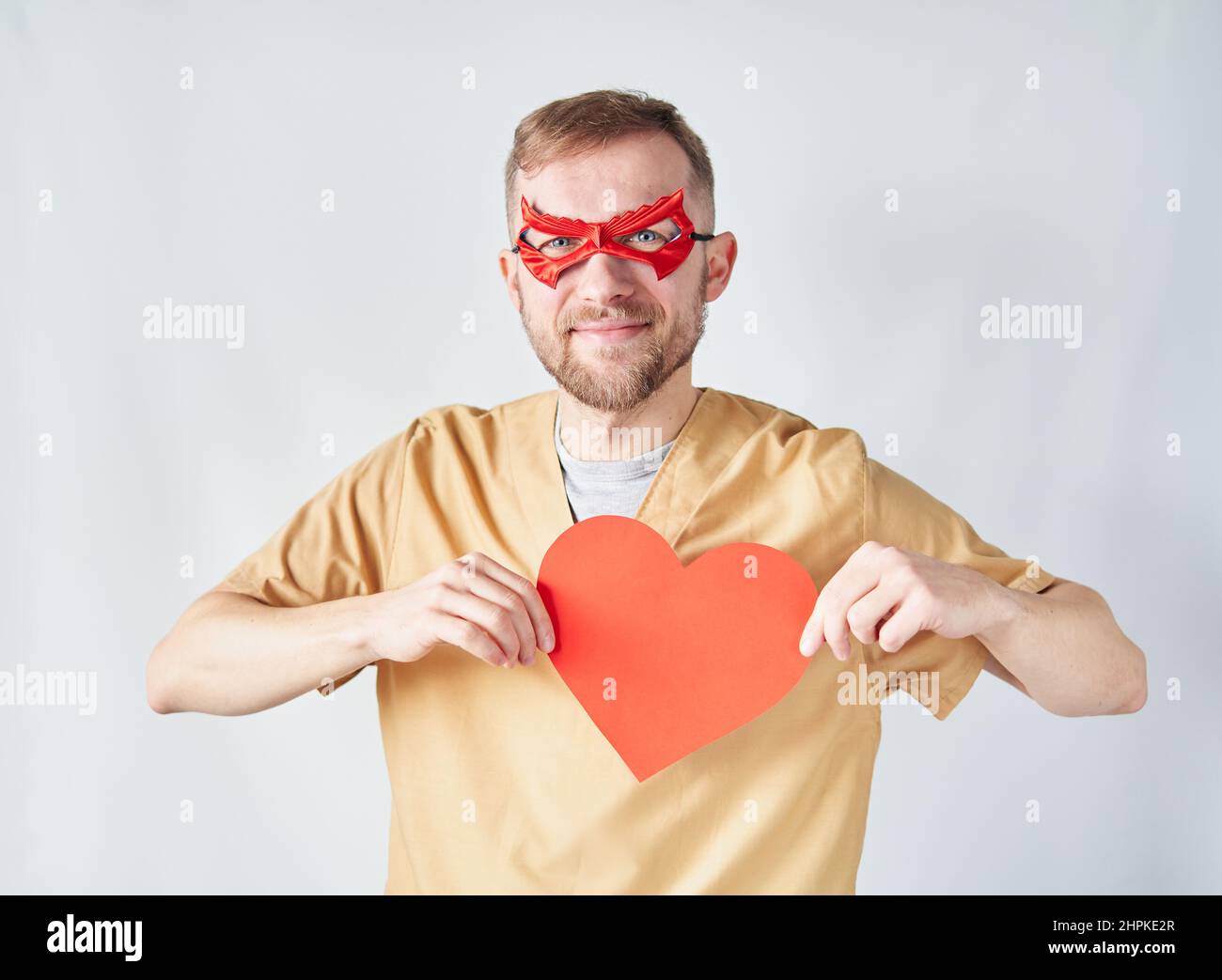 Fröhlich kaukasischen männlichen Kardiologen Arzt in medizinischen Uniform und roten Superhelden Augenmaske mit rotem Papier Herzform. Erwachsene Arzt Chirurg im Krankenhaus posiert über der Wand. Hochwertige Fotos Stockfoto