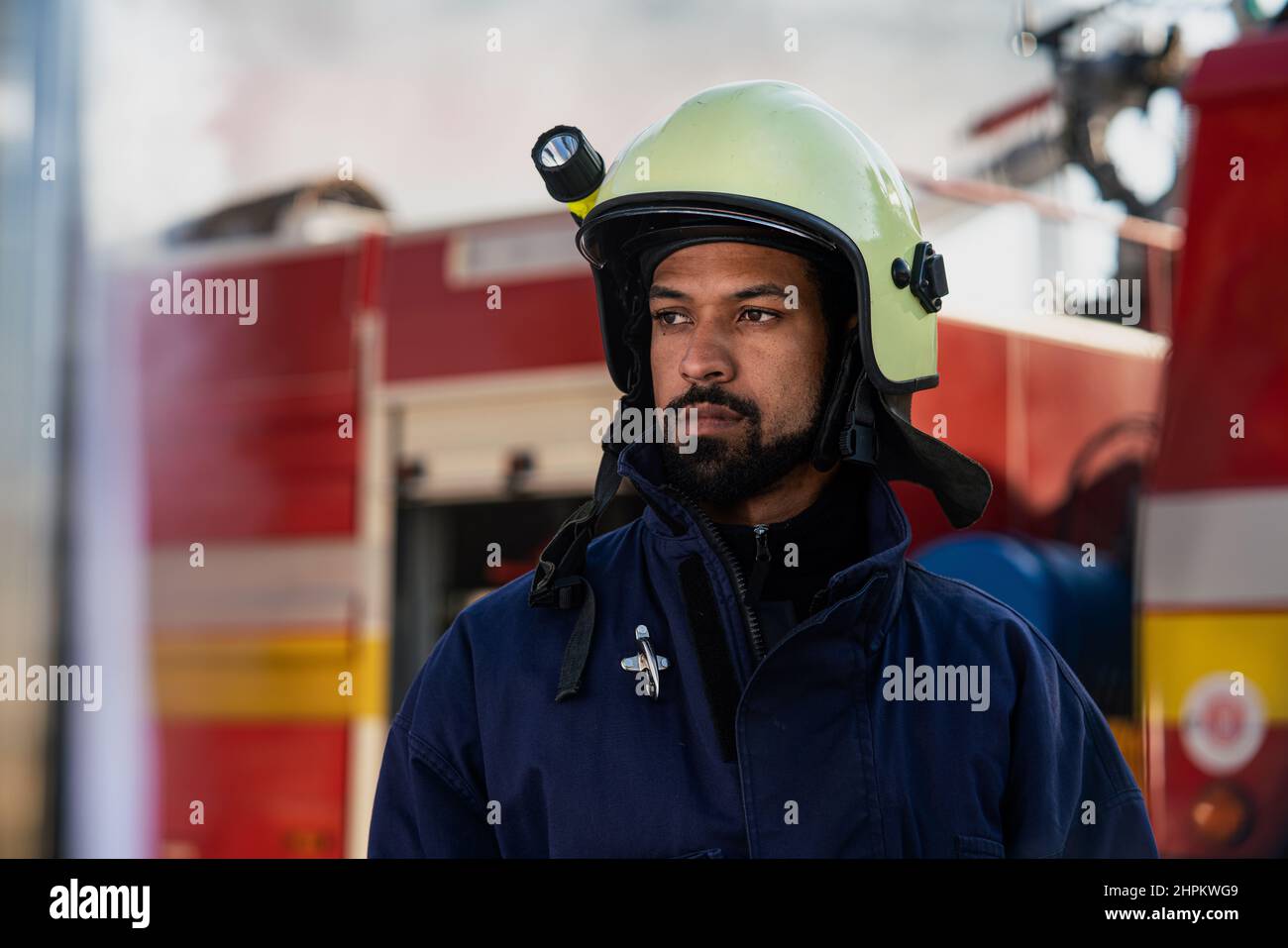 Seriöse junge afroamerikanische Feuerwehrmann mit Feuerwehrauto und Rauch im Hintergrund. Stockfoto