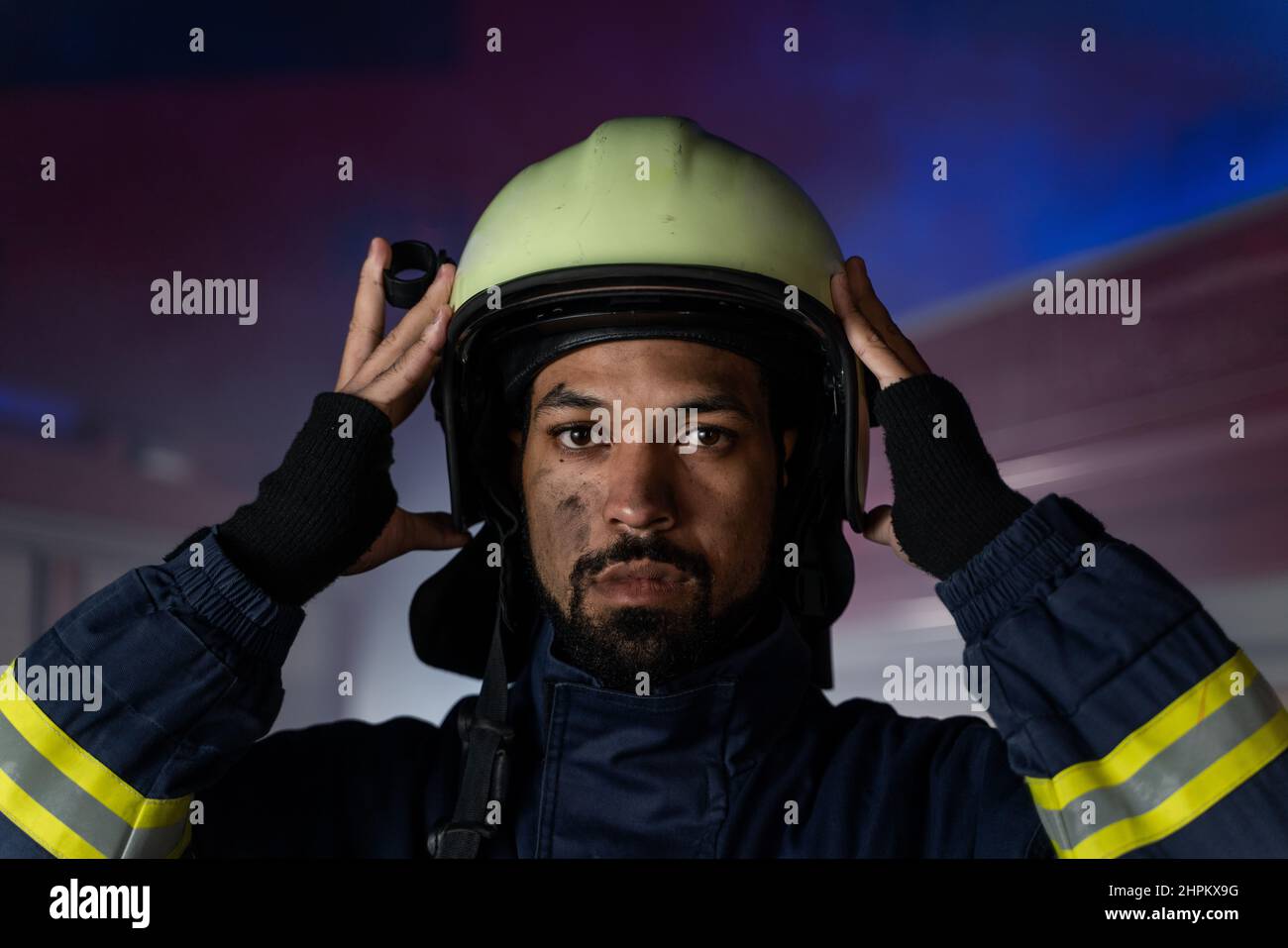 Porträt eines schmutzigen Feuerwehrmanns im Dienst mit Feuerwehrauto im Hintergrund in der Nacht, Blick auf die Kamera. Stockfoto