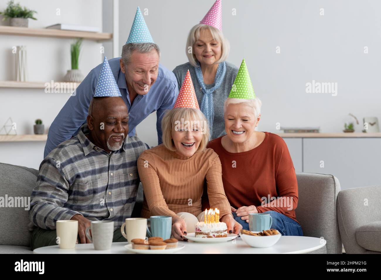 Eine multiethnische Gruppe älterer Menschen, glückliche Männer und Frauen mit bunten Papierhüten, die zu Hause eine Geburtstagsfeier feiern, kaukasische ältere Dame, die eine Geburt hält Stockfoto