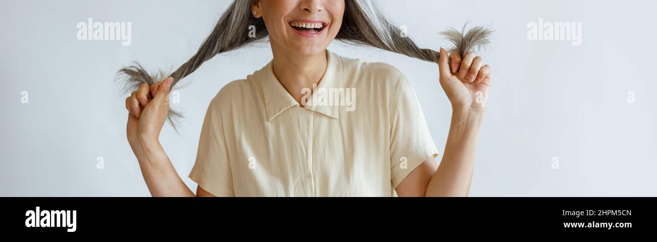 Glückliche asiatische Frau mittleren Alters hält verdrehtes graues Haar auf hellgrauem Hintergrund Stockfoto