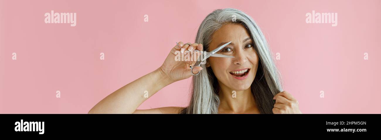 Eine glückliche Frau mittleren Alters mit langen, haarigen Haaren schaut im Studio durch eine offene Schere Stockfoto