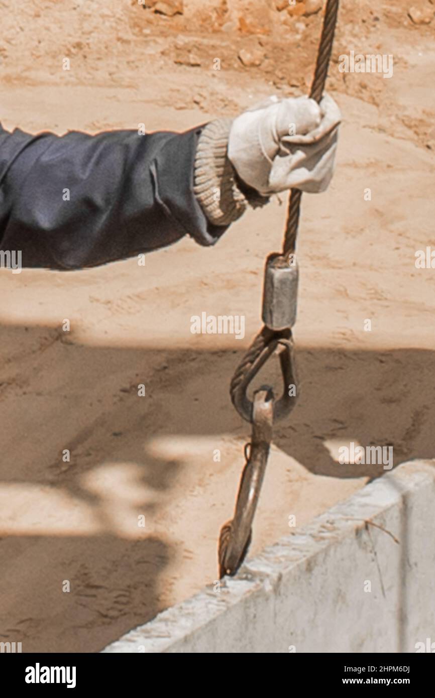 Die Hand des Bauarbeiters männlich nimmt einen schweren Eisenhaken-Hebemechanismus mit einem Stahlseil auf eine Beton-, Zementstruktur. Stockfoto
