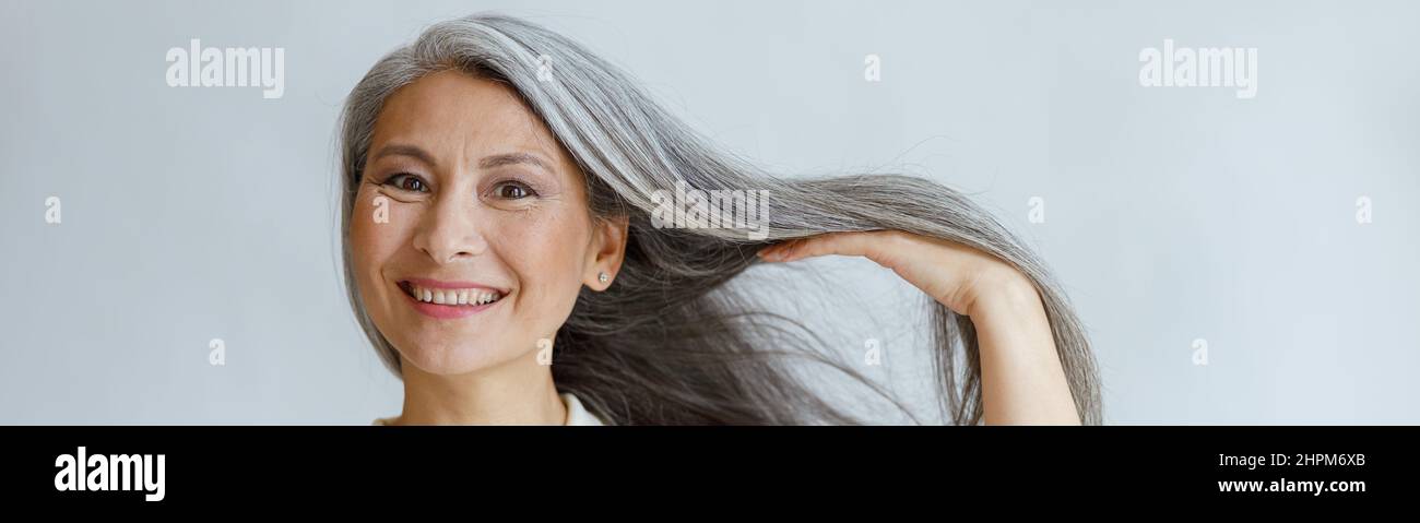 Glückliche asiatische Dame mittleren Alters zeigt natürliches silbernes Haar, das auf hellem Hintergrund posiert Stockfoto