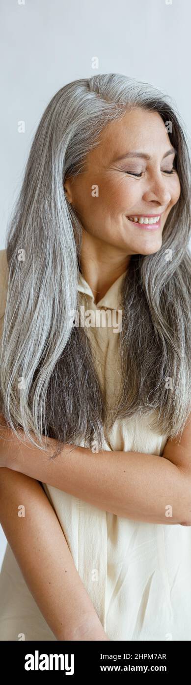 Scheue asiatische Dame mittleren Alters mit langen silbernen Haaren Posen auf hellgrauem Hintergrund Stockfoto