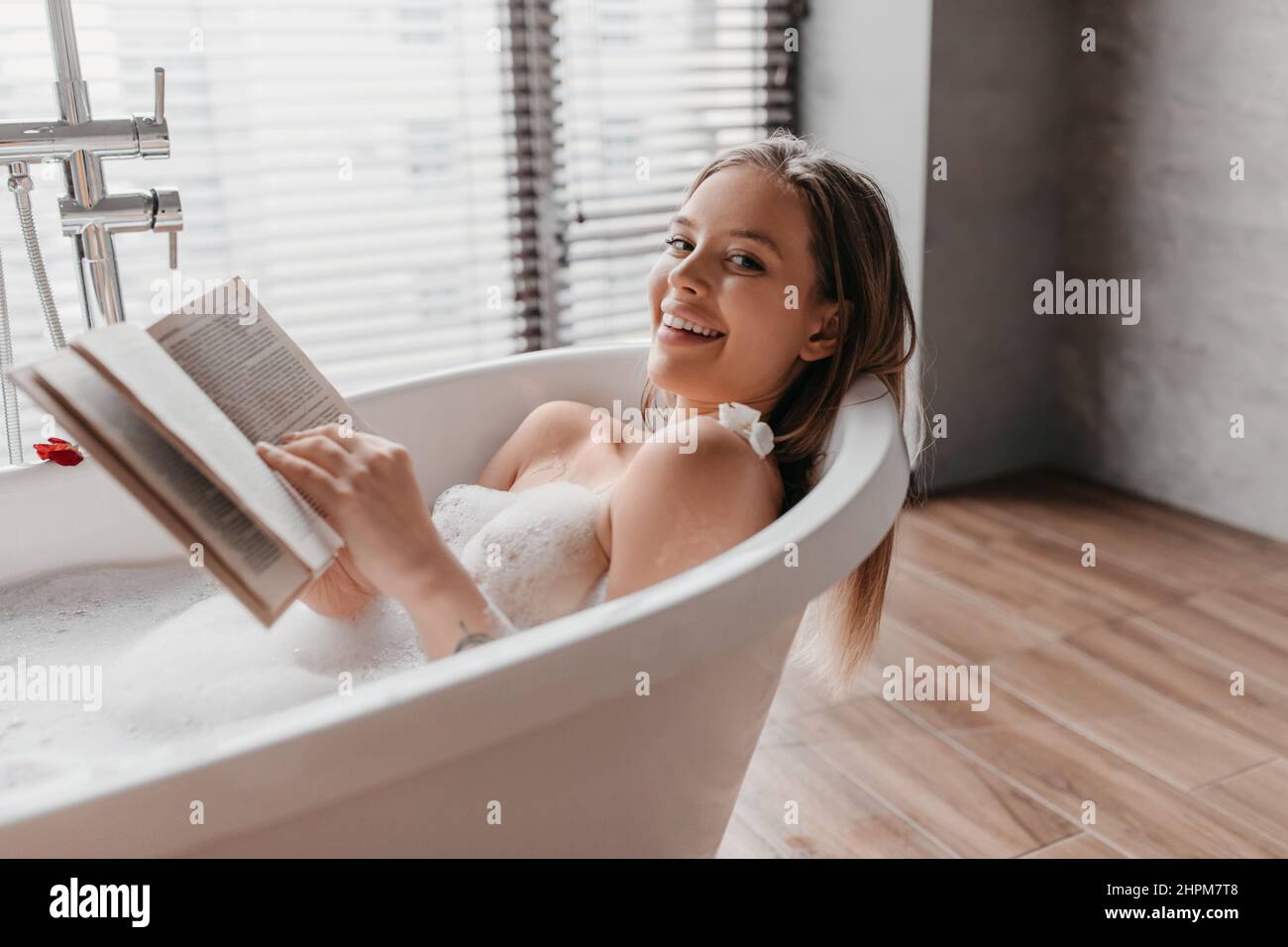 Ruhe im Badezimmer zu Hause. Hübsche junge Frau im Bad mit Schaumstoff, liest Buch und lächelt vor der Kamera Stockfoto
