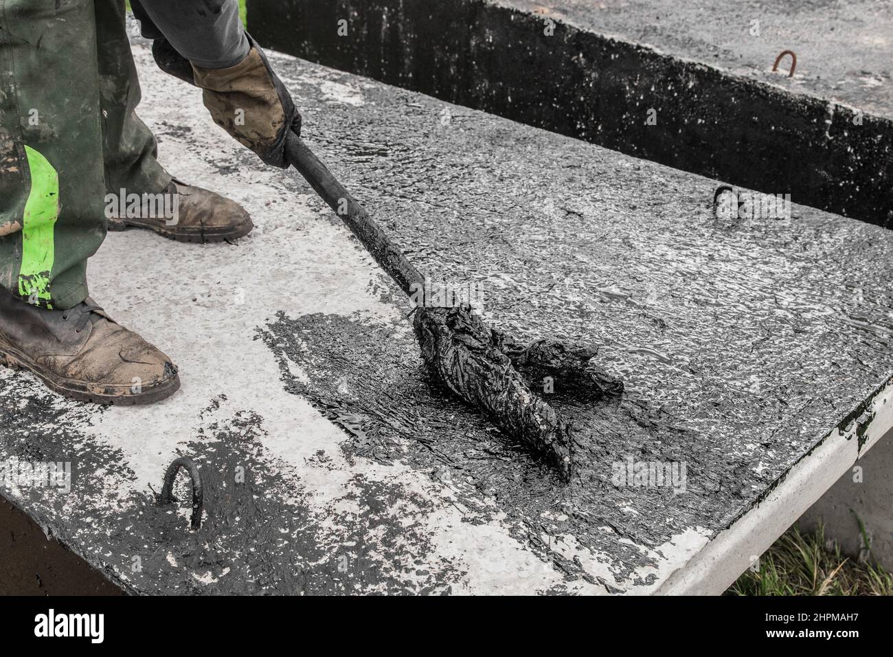 Ein Bauarbeiter ist mit schmutzigen manuellen Arbeiten beschäftigt, um die Betonoberfläche der Struktur zu reparieren und schwarzen Bitumen-Abdichtungsteer aufzutragen. Stockfoto